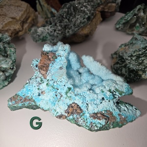 May include: A collection of raw mineral specimens, featuring a prominent piece with a light blue, crystalline structure. The minerals display a mix of colors, including brown, green, and turquoise. The letter "G" is visible in the image.