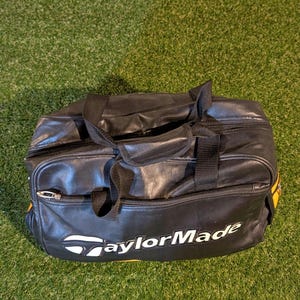 Puede incluir: Una bolsa de deporte negra con el logotipo de TaylorMade en blanco impreso en la parte frontal. La bolsa cuenta con un compartimento principal con cremallera, un bolsillo frontal m&aacute;s peque&ntilde;o con cremallera y dos asas de tela negra.