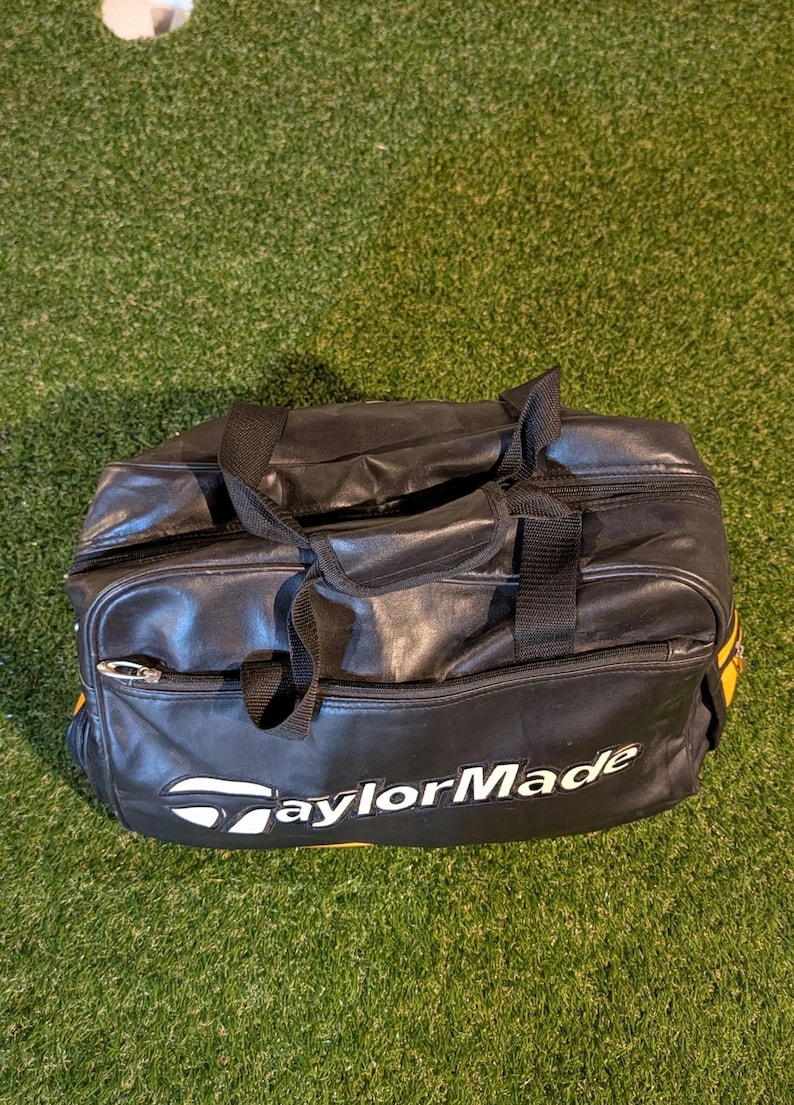 Puede incluir: Una bolsa de deporte negra con el logotipo de TaylorMade en blanco impreso en la parte frontal. La bolsa cuenta con un compartimento principal con cremallera, un bolsillo frontal m&aacute;s peque&ntilde;o con cremallera y dos asas de tela negra.