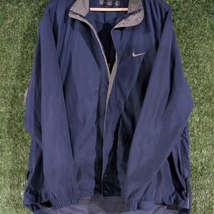 May include: A navy blue Nike windbreaker jacket with a gray collar and a silver Nike swoosh logo. The jacket is hanging on a wooden hanger against a green background.