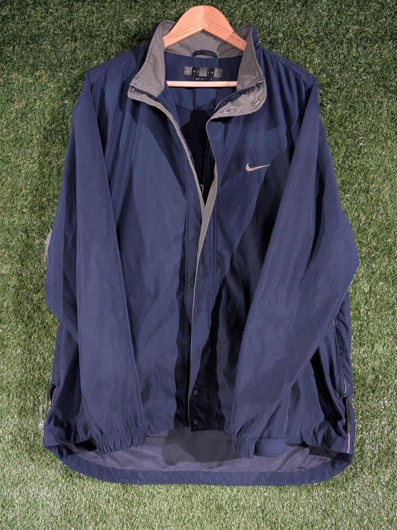 May include: A navy blue Nike windbreaker jacket with a gray collar and a silver Nike swoosh logo. The jacket is hanging on a wooden hanger against a green background.