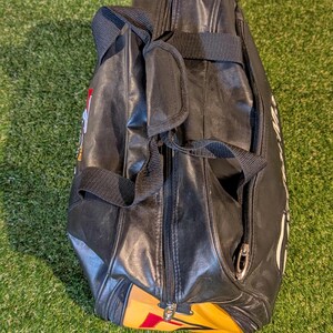 Puede incluir: Una bolsa de deporte negra con acabado sint&eacute;tico liso, que cuenta con un cierre de cremallera central, dos asas de transporte de tela negra con agarre acolchado y un panel de acento amarillo y rojo en la base con letras blancas visibles.