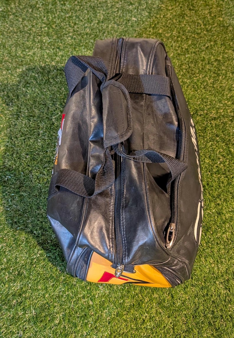 Puede incluir: Una bolsa de deporte negra con acabado sint&eacute;tico liso, que cuenta con un cierre de cremallera central, dos asas de transporte de tela negra con agarre acolchado y un panel de acento amarillo y rojo en la base con letras blancas visibles.