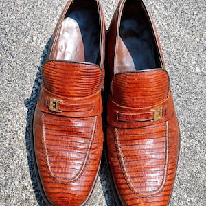 May include: A pair of brown loafers featuring a textured lizard-skin pattern, a rounded toe, and a gold-toned metal hardware accent across the vamp. The interior lining is dark brown, and the shoes have a polished finish with visible stitching.