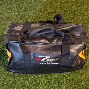 Puede incluir: Una bolsa de deporte rectangular negra con dos asas superiores y cierre de cremallera. La parte frontal presenta el texto en rojo y blanco r7 quad y el logotipo blanco de TaylorMade. Se ven detalles amarillos en los paneles laterales.