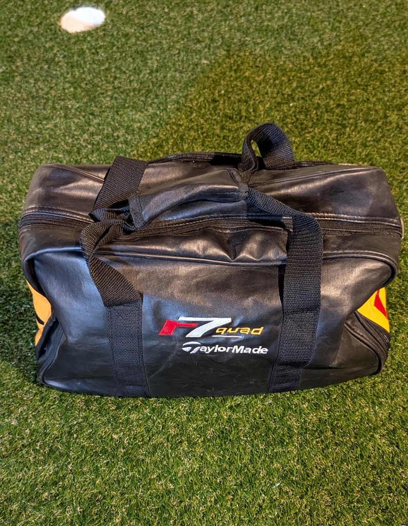Puede incluir: Una bolsa de deporte rectangular negra con dos asas superiores y cierre de cremallera. La parte frontal presenta el texto en rojo y blanco r7 quad y el logotipo blanco de TaylorMade. Se ven detalles amarillos en los paneles laterales.
