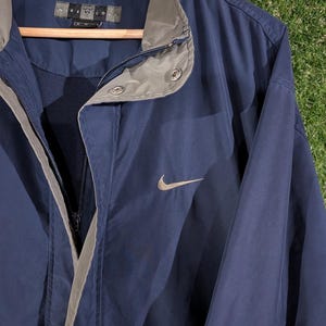 May include: Navy blue Nike golf jacket with a gray collar and trim. The jacket has a full zipper and a small Nike swoosh logo on the chest. The jacket is hanging on a wooden hanger.