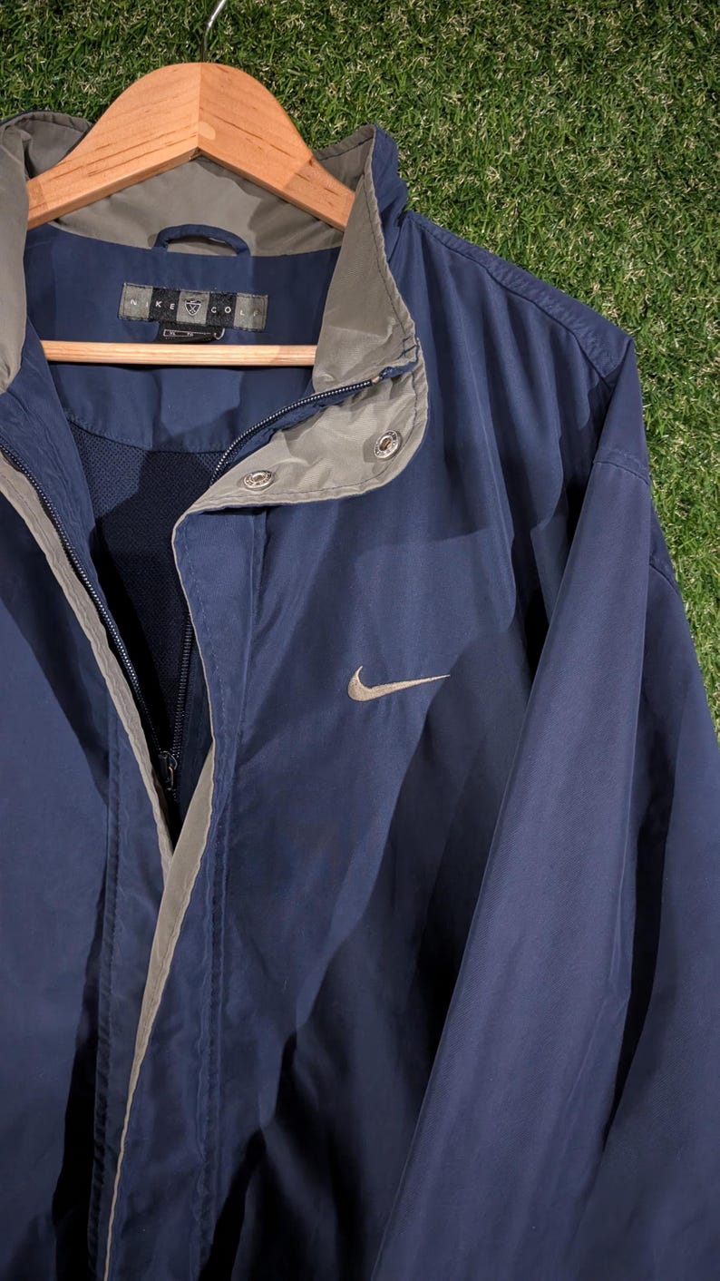 May include: Navy blue Nike golf jacket with a gray collar and trim. The jacket has a full zipper and a small Nike swoosh logo on the chest. The jacket is hanging on a wooden hanger.