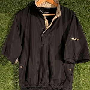 Puede incluir: Chaqueta de golf negra de manga corta con cuello y detalles beige. La chaqueta tiene una botonadura frontal y cintura elástica. La marca "TOP-FLITE" está bordada en dorado en la manga. Colgada en una percha de madera.
