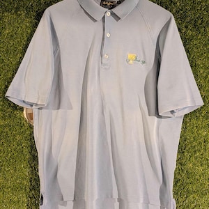 Könnte beinhalten: Hellblaues Poloshirt mit kurzen Ärmeln, Kragen und einer Knopfleiste mit drei Knöpfen. Das Shirt hat ein kleines gesticktes Logo auf der Brust und hängt an einem Holzbügel. Das Shirt ist aus weichem, atmungsaktivem Stoff.