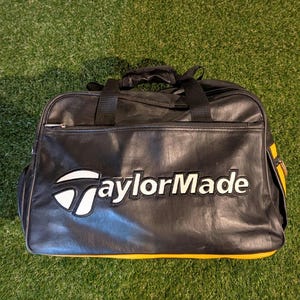 Puede incluir: Una bolsa de deporte negra con detalles en amarillo y el logotipo de TaylorMade bordado en blanco y negro en la parte frontal. La bolsa cuenta con un cierre de cremallera superior, dos asas negras y una construcci&oacute;n de material sint&eacute;tico liso.