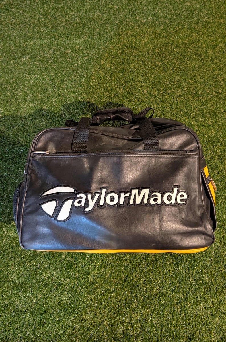 Puede incluir: Una bolsa de deporte negra con detalles en amarillo y el logotipo de TaylorMade bordado en blanco y negro en la parte frontal. La bolsa cuenta con un cierre de cremallera superior, dos asas negras y una construcci&oacute;n de material sint&eacute;tico liso.