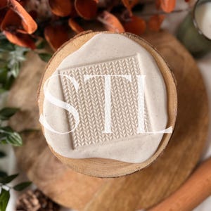 May include: A light-colored, square-shaped stamp with the letters "STI" in a textured, knitted pattern. The stamp is set on a round, wooden base with a light-colored, creamy substance. The image is taken from a top-down perspective.