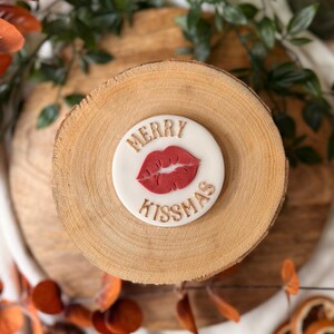 May include: A white circular cookie with the words "MERRY KISS-MAS" in gold lettering and a red lip design. The cookie sits on a wooden slice, surrounded by autumn leaves and greenery.