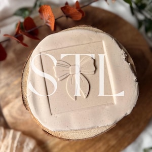 Puede incluir: Un sello beige claro con las letras "STL" y un diseño de lazo. El sello está sobre una base de madera, con un patrón de vetas de madera natural. La imagen está tomada desde arriba.