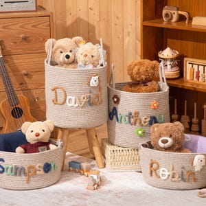 May include: Four beige rope baskets with teddy bears inside. The baskets are labeled "David", "Antheny", "Sampson", and "Robin" in colorful letters. The baskets are on a wooden floor with a ukulele and wooden toys in the background.