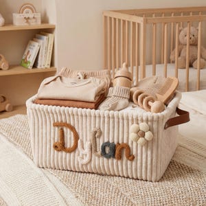 Op de afbeelding: Een crèmekleurige corduroy opbergmand met de naam "Dylan" in bruine, grijze en crèmekleurige letters. De mand bevat babykleding, een houten fles, sokken, een hoed, een kam en een houten speelgoed. Een bloem versiert de zijkant.
