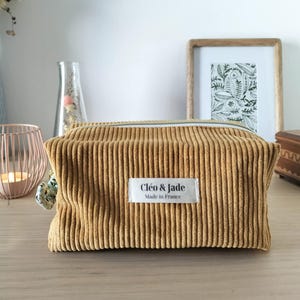 Op de afbeelding: Een rechthoekige make-up tas van goudbruin ribfluweel. De tas heeft een ritssluiting en een label met de tekst "Cléo & Jade, Made in France". De tas staat op een houten oppervlak.