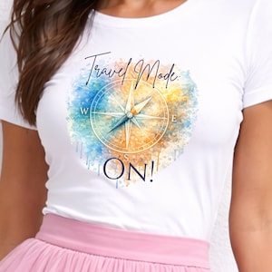 May include: White t-shirt with a watercolour design featuring a compass rose and the words "Travel Mode: ON!" in a script font. The compass is set against a backdrop of blue, orange, and yellow hues.