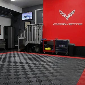 May include: A garage interior with a checkered black and gray floor, red accents, and a red wall featuring the Corvette logo and text. The space includes tool chests, cabinets, and a small staircase.