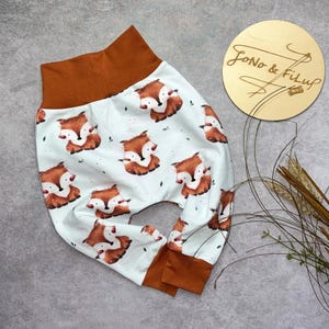 Könnte beinhalten: Babyhose mit einem sich wiederholenden Fuchsmuster auf weißem Grund. Die Bündchen und der Bund sind braun. Eine Holzscheibe mit dem Text "Jono & Filup" ist ebenfalls abgebildet. Das Fuchsmuster ist braun und weiß.
