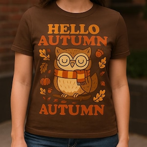 Könnte beinhalten: Braunes T-Shirt mit dem Text "HELLO AUTUMN" in Orange. Das Shirt zeigt eine Cartoon-Eule mit Brille und Schal, umgeben von Herbstblättern und Kürbissen. Das Thema ist der Herbst.