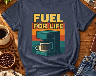 May include: A navy blue t-shirt featuring a graphic print of a teal coffee maker with a white mug. Above the coffee maker, the text reads FUEL FOR LIFE in yellow letters against a background of horizontal orange, yellow, and teal stripes.