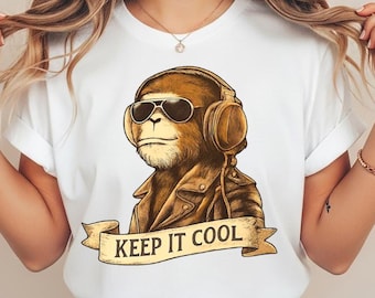 May include: A white t-shirt featuring a graphic of a brown monkey wearing black sunglasses, a leather jacket, and headphones. Below the monkey is a tan banner with the text KEEP IT COOL printed in black capital letters.