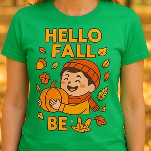 Könnte beinhalten: Ein grünes T-Shirt mit dem Schriftzug "HELLO FALL" in Orange und einer Cartoon-Illustration eines Kindes mit einem Kürbis. Das Design umfasst Herbstblätter und das Wort "BE". Das T-Shirt wird von einer Person getragen, mit einem verschwommenen Herbsthintergrund.
