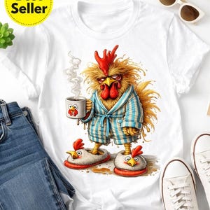 May include: White t-shirt featuring a comical illustration of a disgruntled chicken in a blue and white striped robe, holding a coffee mug. The text "FUNNY CHICKEN T-SHIRT Etsy" is at the top. The shirt is next to blue jeans and white trainers.
