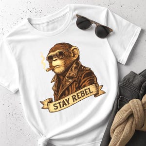 Könnte beinhalten: Weißes T-Shirt mit einem braunen Schimpansen, der eine Sonnenbrille und eine Lederjacke trägt und eine Zigarre raucht. Der Schriftzug "STAY REBEL" ist auf einem Banner darunter aufgedruckt. Eine Sonnenbrille liegt auf dem T-Shirt.