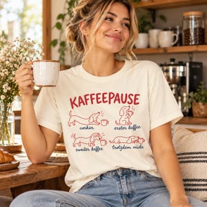 Op de afbeelding: Een crèmekleurig t-shirt met een rood-blauwe afbeelding van vier teckels met de tekst KAFFEEPAUSE, vorher, erster kaffee, zweiter kaffee en trotzdem müde. Het ontwerp toont de honden in verschillende stadia van koffieconsumptie.