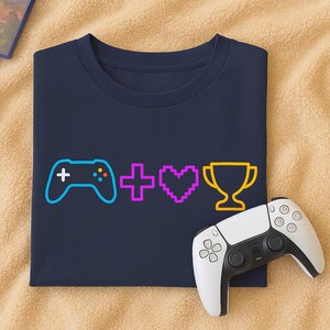Könnte beinhalten: Marineblaues T-Shirt mit einem neongelben und orangefarbenen Videospiel-Controller-Motiv, einem pinkfarbenen Pluszeichen, einem pixeligen lila Herz und einer gelben Trophäe. Ein weißer und schwarzer Videospiel-Controller befindet sich auf der rechten Seite.