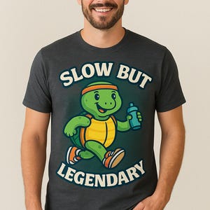 Könnte beinhalten: Dunkelgraues T-Shirt mit einer Cartoon-Schildkröte, die mit einer Wasserflasche rennt. Die Schildkröte ist grün und gelb, trägt ein Stirnband und Turnschuhe. Das Shirt hat die weißen Worte "SLOW BUT LEGENDARY".