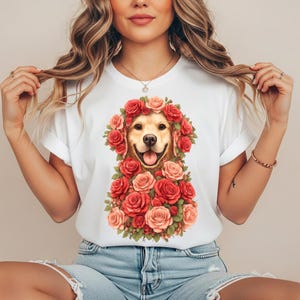 Può includere: Una t-shirt bianca a maniche corte con una grafica centrale di un cane golden retriever circondato da una fitta disposizione di rose rosse e rosa con foglie verdi. Il cane ha un'espressione sorridente con la lingua di fuori.
