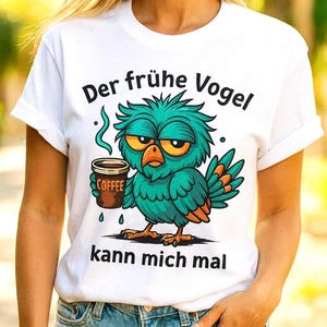 Könnte beinhalten: Weißes T-Shirt mit einem Cartoon-Vogel, der eine Kaffeetasse hält. Der Vogel hat türkisfarbene Federn und orangefarbene Akzente. Der Text "Der frühe Vogel kann mich mal" ist aufgedruckt.