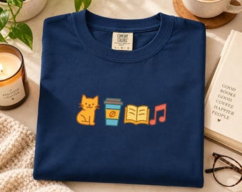 May include: A navy blue t-shirt featuring a graphic print of an orange cat, a blue coffee cup, an open yellow book, and a red musical note. The shirt has a Comfort Colors brand label at the neck with the text L G G printed on the tag.
