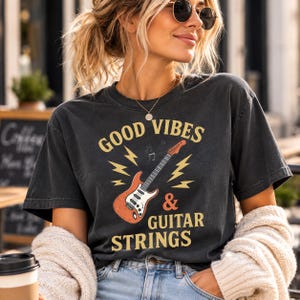Könnte beinhalten: Ein anthrazitfarbenes Kurzarm-T-Shirt mit einer Grafik einer orangefarbenen E-Gitarre, gelben Blitzen und dem Text "GOOD VIBES & GUITAR STRINGS" in einer fetten Vintage-Schriftart, die mittig auf der Vorderseite des Kleidungsstücks platziert ist.