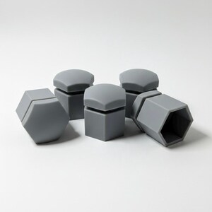 May include: A collection of six gray, geometric bottle stoppers. The stoppers have a square base and a rounded, hexagonal top. One stopper is lying on its side, revealing its hollow interior. The stoppers are made of a soft, matte material.