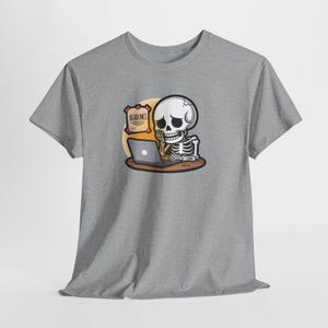 May include: A gray t-shirt featuring a cartoon skeleton working on a laptop. The skeleton is sitting at a desk with a sign that reads "DEADLINES". The design is in shades of white, black, and brown.