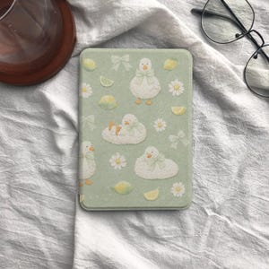 May include: A rectangular, light green case featuring a pattern of white ducks with green bows, daisies, lemon slices, and bows. The case has a rounded edge and a smooth surface. The overall design is whimsical and pastel-colored.
