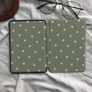 Puede incluir: Una funda para lector electr&oacute;nico de color verde salvia con un patr&oacute;n repetitivo de peque&ntilde;as flores blancas de seis p&eacute;talos. La funda est&aacute; abierta, revelando una superficie interior lisa con el mismo dise&ntilde;o floral, junto a unas gafas negras.
