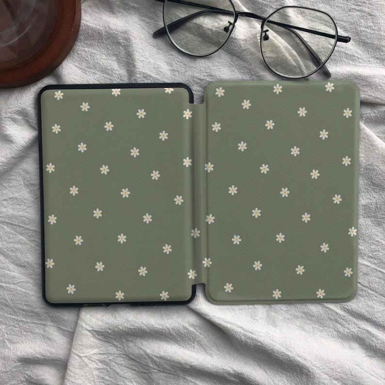 Puede incluir: Una funda para lector electr&oacute;nico de color verde salvia con un patr&oacute;n repetitivo de peque&ntilde;as flores blancas de seis p&eacute;talos. La funda est&aacute; abierta, revelando una superficie interior lisa con el mismo dise&ntilde;o floral, junto a unas gafas negras.