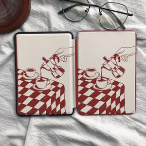 May include: A protective e-reader cover featuring a red and white checkered tablecloth pattern. The design displays a hand pouring coffee from a moka pot into a cup, with a matching saucer and cup set resting on the table surface.
