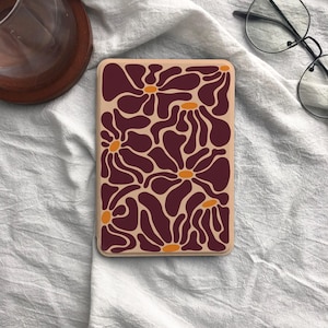 Puede incluir: Una funda rectangular para teléfono con un estampado floral. El diseño presenta flores de color burdeos oscuro con centros naranjas sobre un fondo beige. La funda probablemente está hecha de plástico duro.