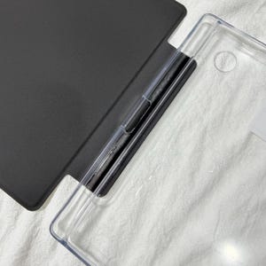 May include: A protective tablet case featuring a solid matte black cover attached to a transparent, rigid plastic shell. The design includes a dedicated vertical slot for a stylus pen along the inner spine, with a circular cutout for a camera lens.