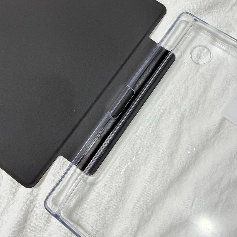 May include: A protective tablet case featuring a solid matte black cover attached to a transparent, rigid plastic shell. The design includes a dedicated vertical slot for a stylus pen along the inner spine, with a circular cutout for a camera lens.