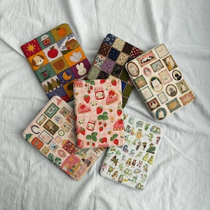 May include: A collection of fabric-covered book covers with various patterns. Designs include patchwork, strawberries, framed cat portraits, and whimsical illustrations of animals and food. The covers are arranged on a white surface.