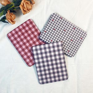 May include: Three square coasters with a gingham pattern in shades of red, gray, and purple. The red coaster has a solid red and pink gingham pattern. The gray coaster has a gray and white gingham pattern with small red heart accents.