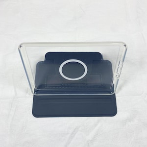 Puede incluir: Una funda protectora transparente para tableta con una base de soporte plegable en color azul oscuro. La parte posterior incluye un anillo de carga magn&eacute;tico circular centrado en un panel azul oscuro, con recortes para botones laterales.